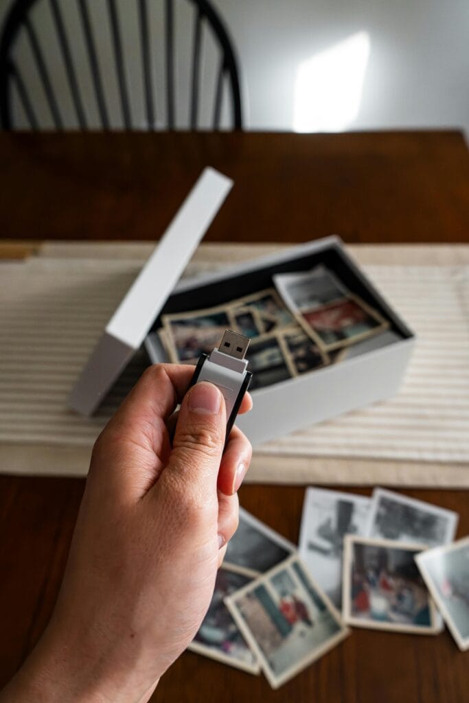 pexels-photo-34384371-34384371 Hand holding USB drive above box of old family photos on a table.