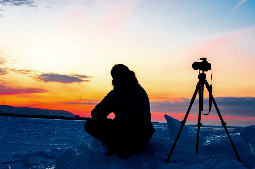 A photographer sits next to a camera on a tripod against a vibrant icy sunset.